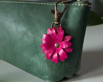 May include: A dark green leather pouch with a brass zipper and a pink leather flower charm. The flower charm is attached with a brass clasp. The pouch is a solid color and has a simple design.
