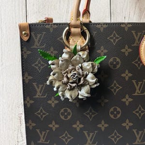 May include: A brown leather handbag with a gold-tone chain strap and a beige flower charm attached to the handle. The bag has a brown and beige monogram pattern.