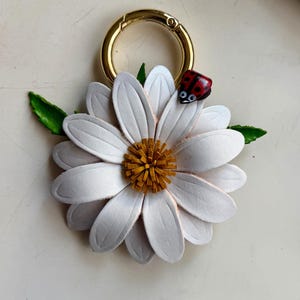 May include: A white daisy-shaped keychain with a gold-colored clasp. The flower has a yellow center and a small red ladybug on one petal. Green leaves are attached to the flower.