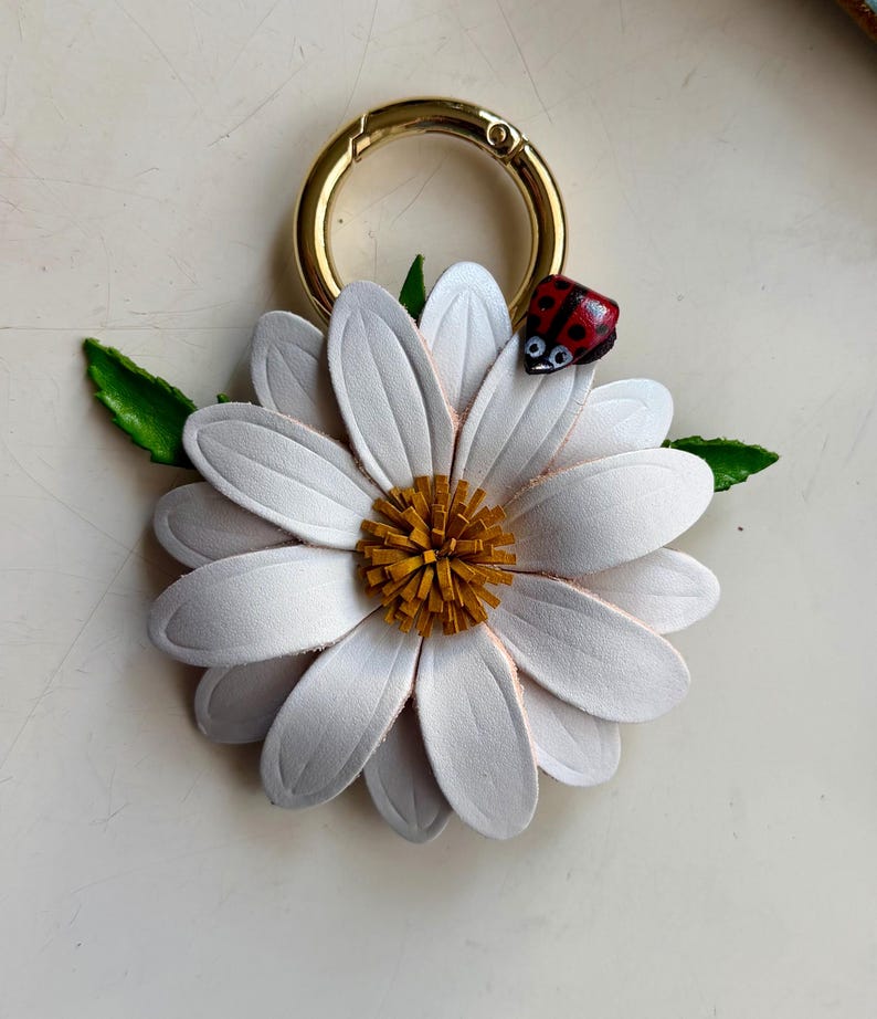 May include: A white daisy-shaped keychain with a gold-colored clasp. The flower has a yellow center and a small red ladybug on one petal. Green leaves are attached to the flower.