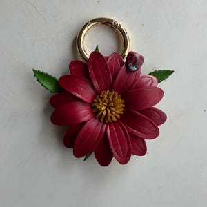 May include: A burgundy leather flower keychain with a gold-colored clasp. The flower has a yellow center and green leaves. A small ladybug is perched on one of the petals. The keychain is a decorative accessory.