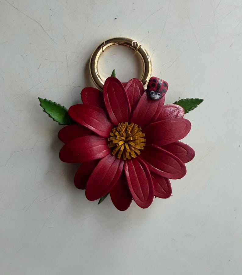 May include: A burgundy leather flower keychain with a gold-colored clasp. The flower has a yellow center and green leaves. A small ladybug is perched on one of the petals. The keychain is a decorative accessory.