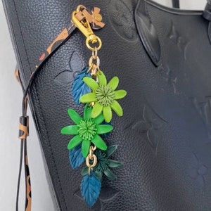 K&ouml;nnte beinhalten: Schwarze Ledertasche mit gepr&auml;gtem Blumenmuster. Ein goldfarbener Anh&auml;nger mit gr&uuml;nen und blauen Blumenakzenten h&auml;ngt am Griff. Die Tasche hat einen Leopardenmusterriemen.
