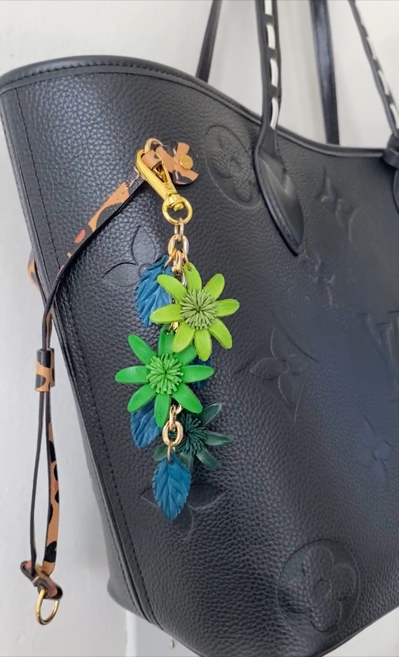 K&ouml;nnte beinhalten: Schwarze Ledertasche mit gepr&auml;gtem Blumenmuster. Ein goldfarbener Anh&auml;nger mit gr&uuml;nen und blauen Blumenakzenten h&auml;ngt am Griff. Die Tasche hat einen Leopardenmusterriemen.