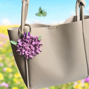 Op de afbeelding: Een taupekleurige tote bag met een textuur, versierd met een paarse bloemenbedel. De tas heeft een eenvoudig ontwerp met twee handvatten en een gouden ring. Vlinders fladderen op de achtergrond tegen een blauwe lucht en een bloemenveld.