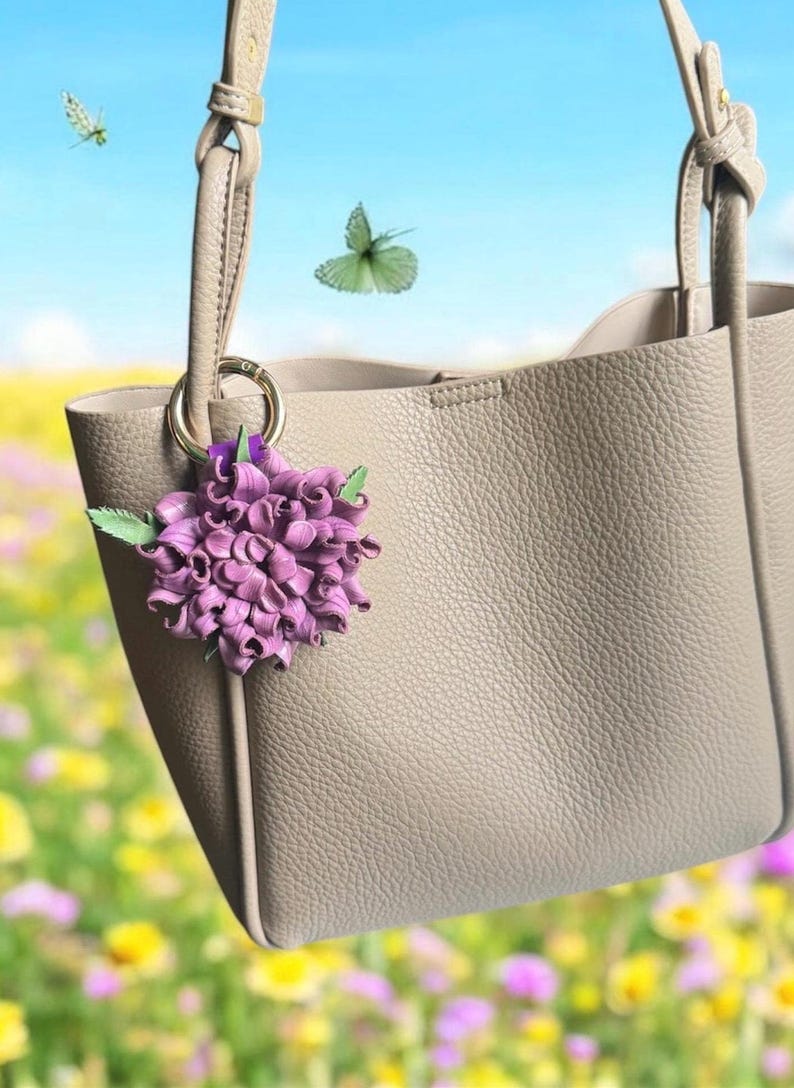 Op de afbeelding: Een taupekleurige tote bag met een textuur, versierd met een paarse bloemenbedel. De tas heeft een eenvoudig ontwerp met twee handvatten en een gouden ring. Vlinders fladderen op de achtergrond tegen een blauwe lucht en een bloemenveld.