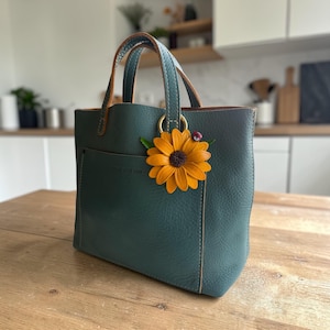 May include: A teal leather tote bag with a sunflower accent. The bag has a front pocket and brown handles. A decorative sunflower with a ladybug charm is attached to the handle ring. The bag is sitting on a wooden table.