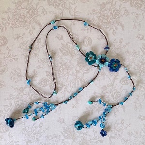 May include: A long brown cord necklace featuring three central layered flower charms in shades of blue and teal. The cord is accented with small light blue and turquoise beads, ending in two dangling sections with additional blue flower charms.
