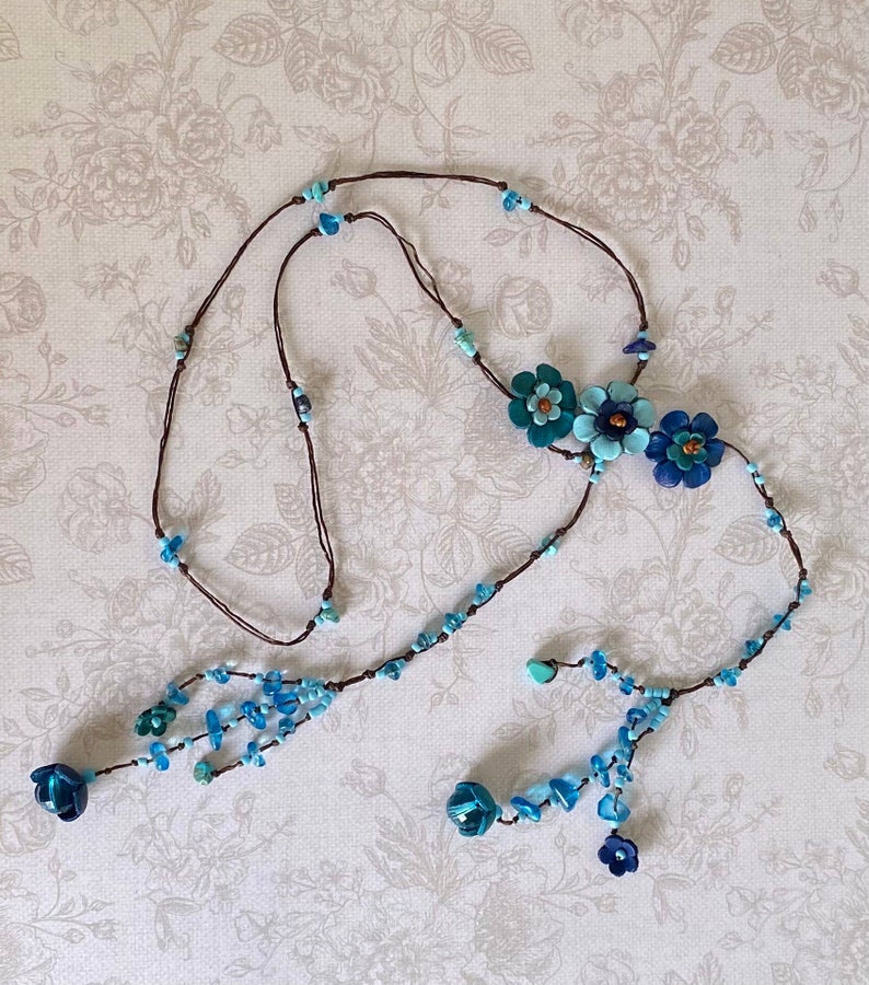 May include: A long brown cord necklace featuring three central layered flower charms in shades of blue and teal. The cord is accented with small light blue and turquoise beads, ending in two dangling sections with additional blue flower charms.