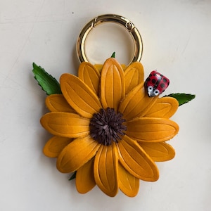 May include: A handcrafted leather sunflower keychain with a gold-colored clasp. The sunflower has yellow petals and a dark brown center. A small ladybug with red and black spots is perched on a green leaf.