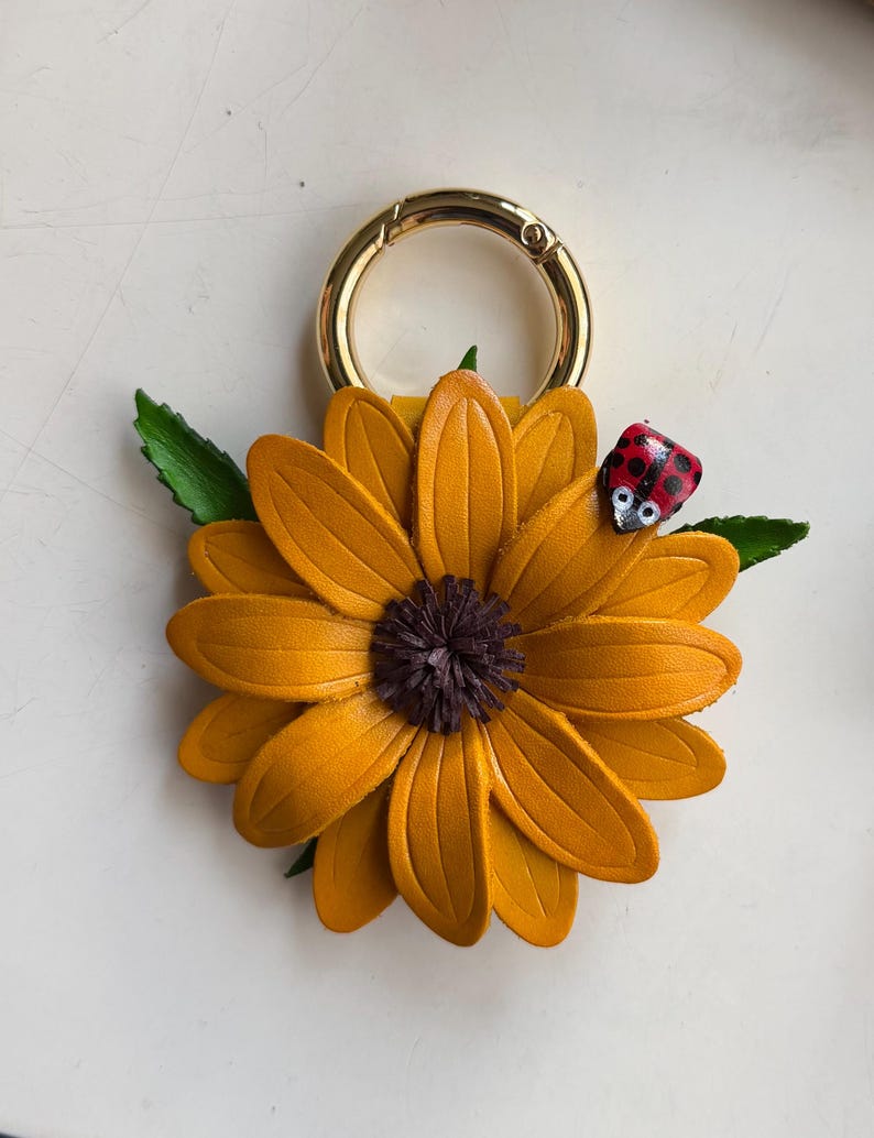May include: A handcrafted leather sunflower keychain with a gold-colored clasp. The sunflower has yellow petals and a dark brown center. A small ladybug with red and black spots is perched on a green leaf.