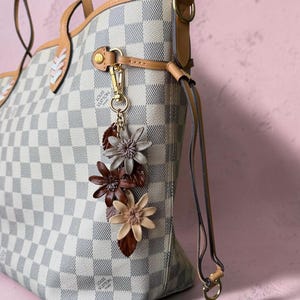 May include: A white and gray checkered tote bag with tan leather trim and handles. A gold-tone metal clasp attaches a decorative charm featuring three layered leather flowers in shades of beige, brown, and dark gray with matching leaves.