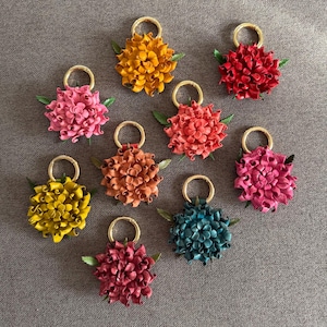 Op de afbeelding: Een verzameling handgemaakte leren bloemvormige sleutelhangers in verschillende kleuren, waaronder roze, geel, oranje, rood en blauwgroen. Elke sleutelhanger heeft een goudkleurige ring en kleine groene blaadjes, op een grijze ondergrond.