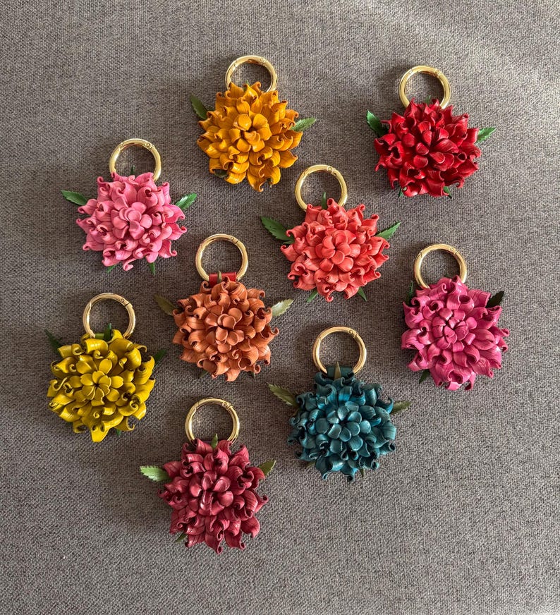 Op de afbeelding: Een verzameling handgemaakte leren bloemvormige sleutelhangers in verschillende kleuren, waaronder roze, geel, oranje, rood en blauwgroen. Elke sleutelhanger heeft een goudkleurige ring en kleine groene blaadjes, op een grijze ondergrond.