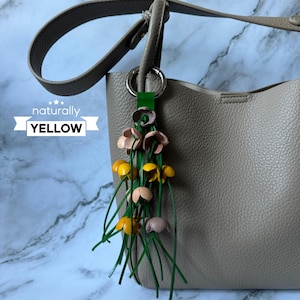 K&ouml;nnte beinhalten: Eine beige Lederhandtasche mit einem gelben, rosa und gr&uuml;nen Lederblumen-Anh&auml;nger, der am Henkel befestigt ist. Der Anh&auml;nger tr&auml;gt die Aufschrift "naturally YELLOW" auf einem wei&szlig;en Band.