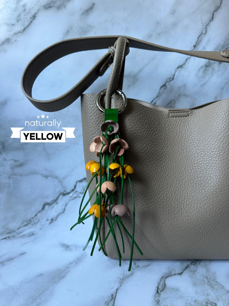 K&ouml;nnte beinhalten: Eine beige Lederhandtasche mit einem gelben, rosa und gr&uuml;nen Lederblumen-Anh&auml;nger, der am Henkel befestigt ist. Der Anh&auml;nger tr&auml;gt die Aufschrift "naturally YELLOW" auf einem wei&szlig;en Band.