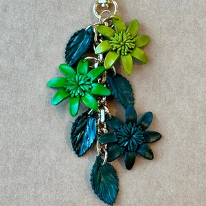 K&ouml;nnte beinhalten: Ein floraler Schl&uuml;sselanh&auml;nger mit einem goldfarbenen Verschluss und einer Kette. Der Schl&uuml;sselanh&auml;nger zeigt drei Bl&uuml;ten in Gr&uuml;n- und Dunkelgr&uuml;nt&ouml;nen mit passenden gr&uuml;nen Bl&auml;ttern. Das Wort "Greenery" ist oben aufgedruckt.