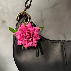 May include: A black leather handbag with a pink flower charm attached. The flower is a vibrant pink with green leaves, and the bag has a gold-coloured ring and strap.