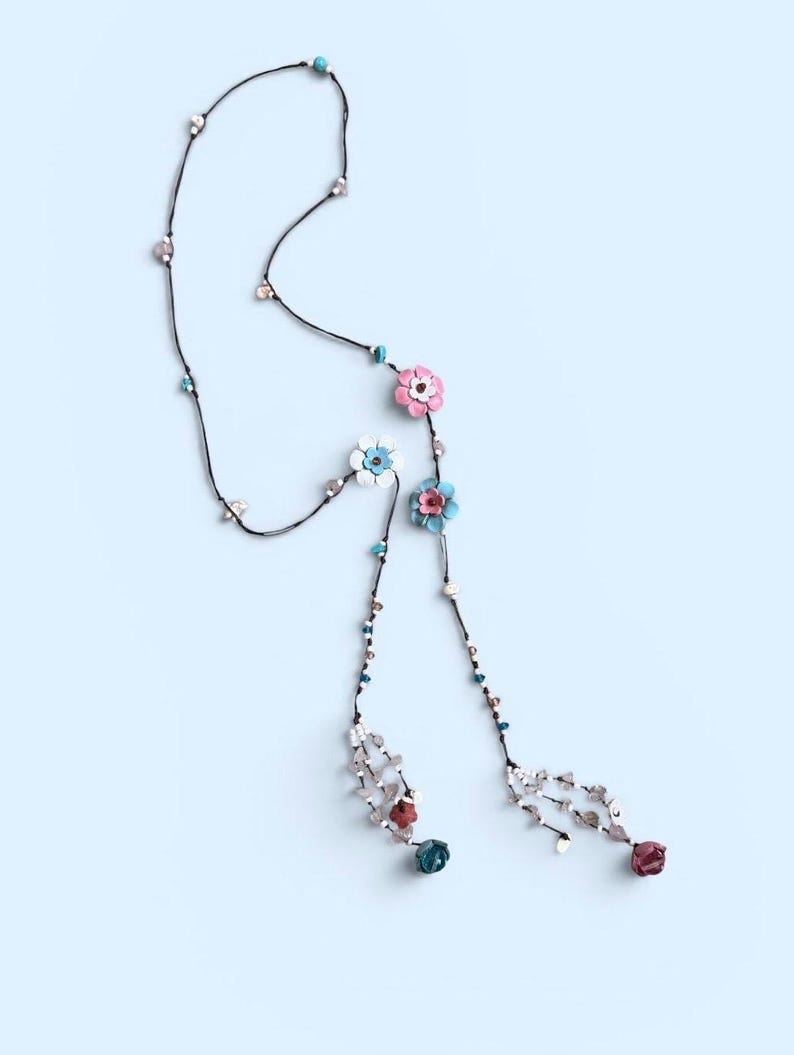 May include: A long lariat-style necklace featuring a thin black cord adorned with small turquoise, clear, and metallic beads. The necklace includes several multi-colored flower charms in shades of pink, white, light blue, and dark red, ending in tassels.