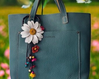 Könnte beinhalten: Türkisfarbene Ledertasche mit einem dekorativen Gänseblümchen- und Blumenanhänger. Die Tasche hat eine Tasche auf der Vorderseite und braune Ledergriffe. Der Anhänger zeigt eine große weiße Gänseblümchen mit gelbem Zentrum und kleinen bunten Blumen.