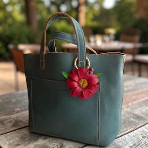 May include: A teal leather tote bag with a red flower and ladybug charm. The bag has a front pocket, a gold ring detail, and brown handles. The bag is sitting on a wooden surface.