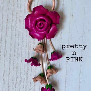 May include: A pink leather rose with a loop at the top and a dangling chain of smaller pink and beige leather roses. The text "pretty in PINK" is written below the roses.