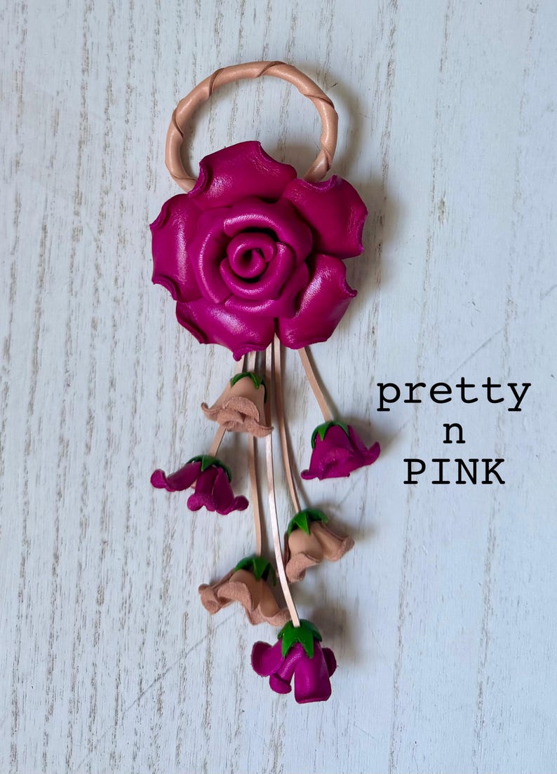 May include: A pink leather rose with a loop at the top and a dangling chain of smaller pink and beige leather roses. The text "pretty in PINK" is written below the roses.