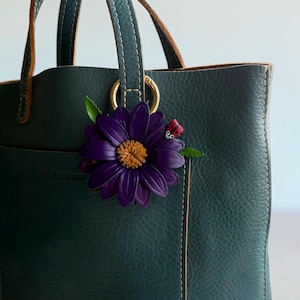 May include: A dark teal leather tote bag with brown handles and gold hardware. A large purple flower with a yellow center and a small ladybug accent the bag's exterior. The bag has a pocket on the front.