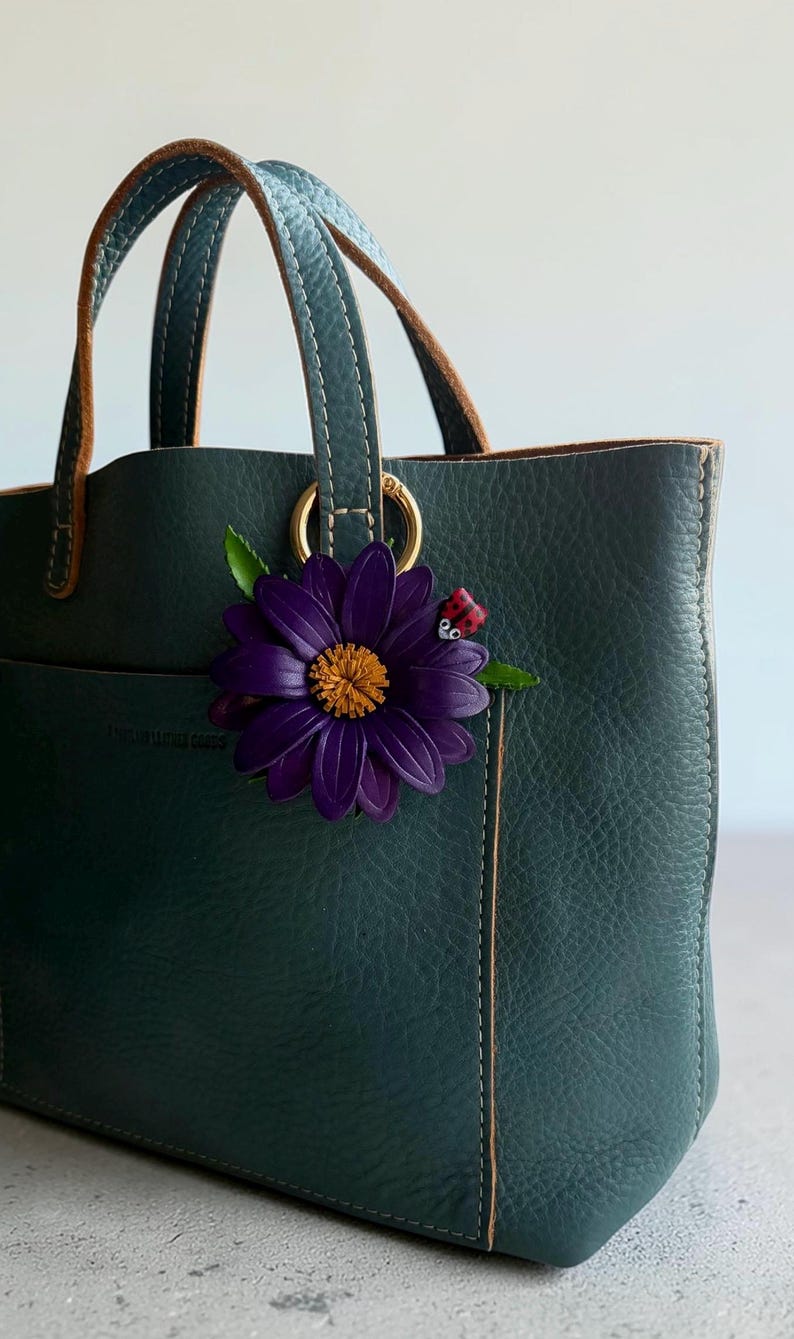 May include: A dark teal leather tote bag with brown handles and gold hardware. A large purple flower with a yellow center and a small ladybug accent the bag's exterior. The bag has a pocket on the front.