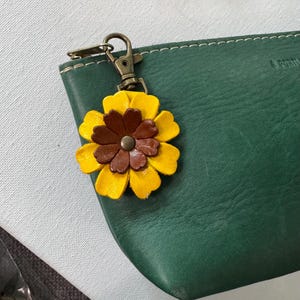 May include: A yellow and brown leather flower charm with a bronze clasp attached to a dark green leather pouch. The flower has multiple layers of petals and a central bronze button. The pouch has white stitching.