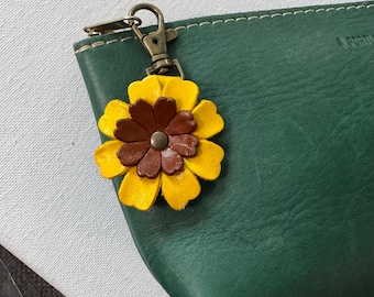 May include: A yellow and brown leather flower charm with a bronze clasp attached to a dark green leather pouch. The flower has multiple layers of petals and a central bronze button. The pouch has white stitching.