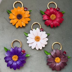 May include: Five flower-shaped keychains in shades of orange, red, white, purple, and brown. Each keychain features a gold ring, green leaves, and a small ladybug. The flower petals are detailed, and the center of each flower is a contrasting color.