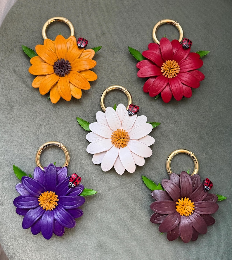 May include: Five flower-shaped keychains in shades of orange, red, white, purple, and brown. Each keychain features a gold ring, green leaves, and a small ladybug. The flower petals are detailed, and the center of each flower is a contrasting color.