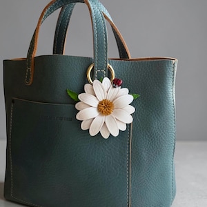 May include: A teal leather tote bag with a daisy and ladybug charm. The bag has a front pocket and brown handles. The daisy charm is white with a yellow center. The ladybug is red with black spots.