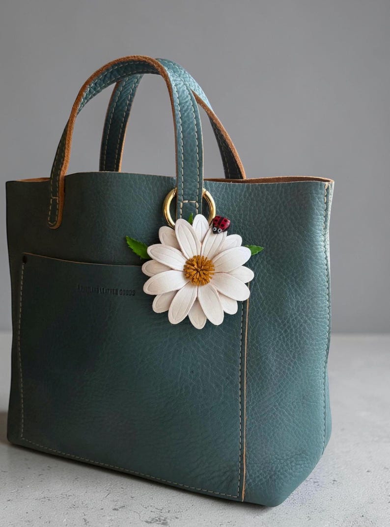 May include: A teal leather tote bag with a daisy and ladybug charm. The bag has a front pocket and brown handles. The daisy charm is white with a yellow center. The ladybug is red with black spots.