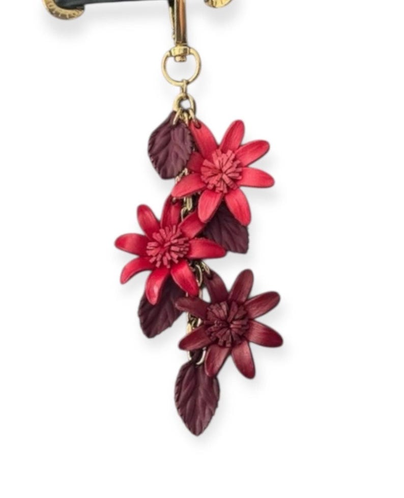 K&ouml;nnte beinhalten: Ein Schl&uuml;sselanh&auml;nger mit floralem Thema mit drei roten Blumenanh&auml;ngern mit detaillierten Bl&uuml;tenbl&auml;ttern und dunkelroten Bl&auml;ttern. Die Blumen sind durch eine goldene Kette verbunden und an einem goldfarbenen Verschluss befestigt.
