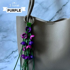 K&ouml;nnte beinhalten: Eine hellgraue Ledertasche mit einem lila, pink und gr&uuml;nen Lederblumen-Anh&auml;nger am Henkel. Der Anh&auml;nger tr&auml;gt die Aufschrift "3 tone PURPLE" auf einem wei&szlig;en Band.
