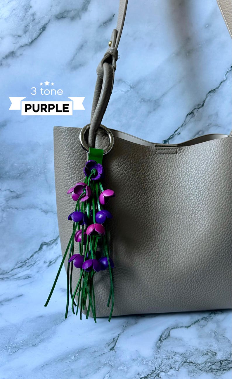 K&ouml;nnte beinhalten: Eine hellgraue Ledertasche mit einem lila, pink und gr&uuml;nen Lederblumen-Anh&auml;nger am Henkel. Der Anh&auml;nger tr&auml;gt die Aufschrift "3 tone PURPLE" auf einem wei&szlig;en Band.