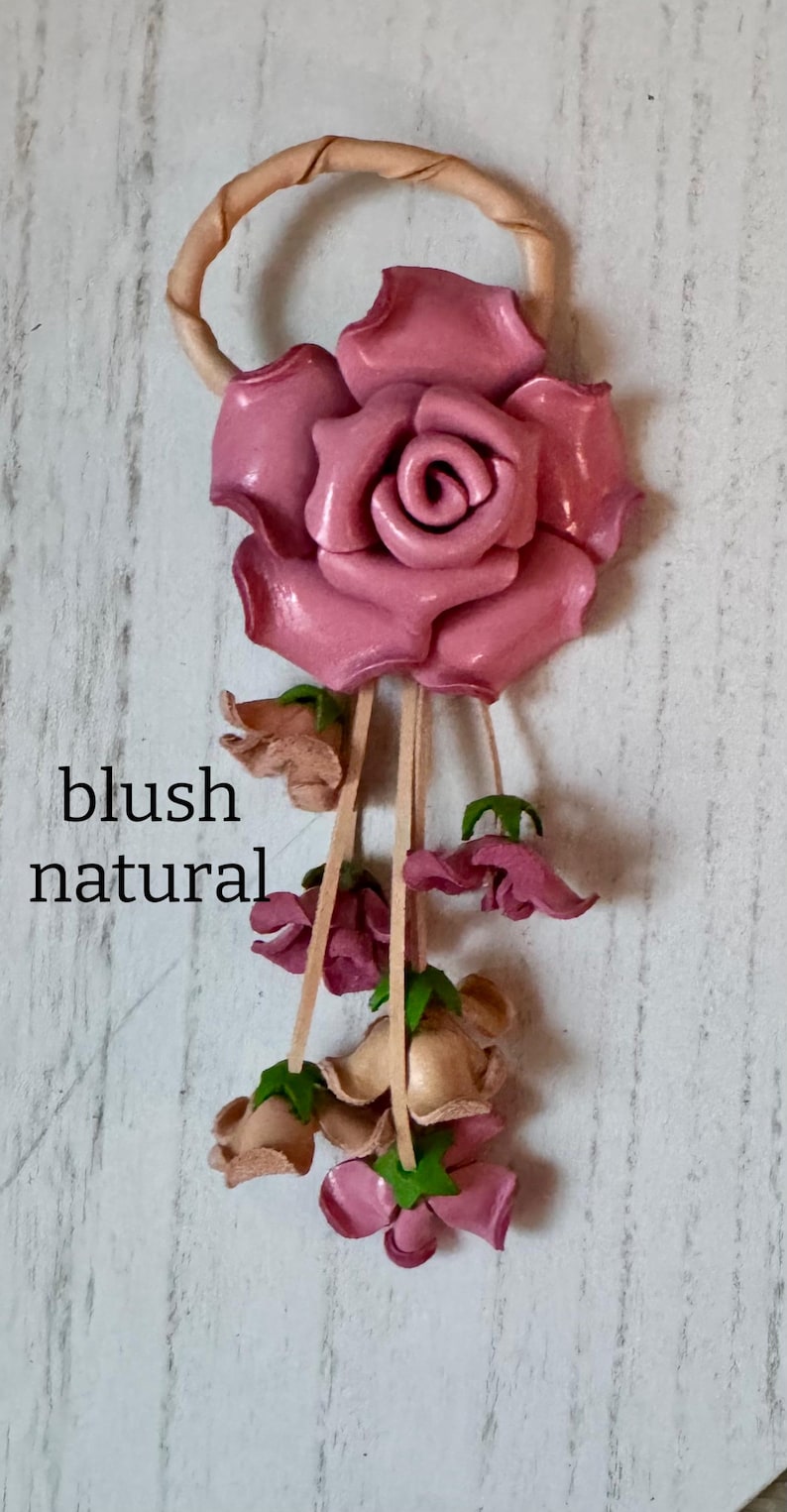 May include: A leather rose keychain with a blush pink rose and smaller pink and beige flowers hanging from a tan leather loop.