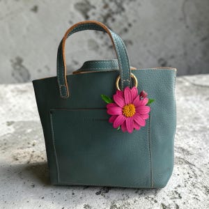 May include: A teal leather tote bag with a small front pocket and a top handle. A pink flower with a yellow center and a small ladybug charm hangs from a gold ring on the handle. The bag has a structured shape.
