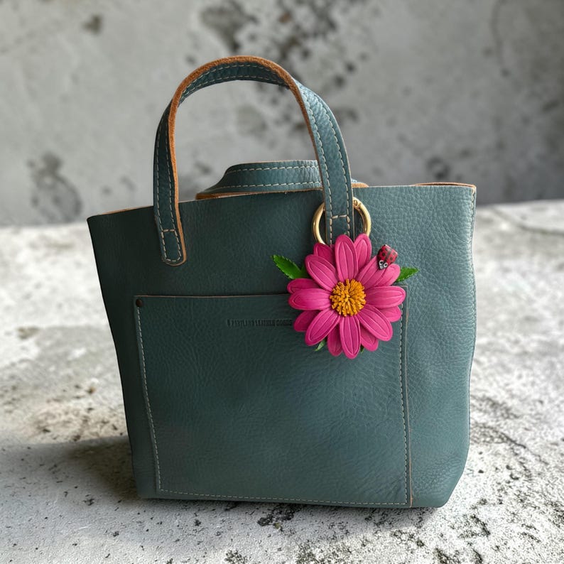 May include: A teal leather tote bag with a small front pocket and a top handle. A pink flower with a yellow center and a small ladybug charm hangs from a gold ring on the handle. The bag has a structured shape.