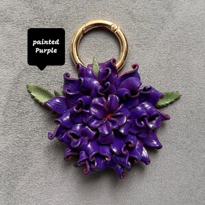 Op de afbeelding: Een paarse bloemvormige sleutelhanger met een goudkleurige ring sluiting. De bloem heeft gedetailleerde bloemblaadjes en groene bladeren. De tekst "painted Purple" staat in een zwarte box.