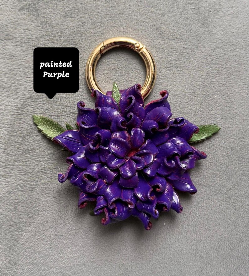 Op de afbeelding: Een paarse bloemvormige sleutelhanger met een goudkleurige ring sluiting. De bloem heeft gedetailleerde bloemblaadjes en groene bladeren. De tekst "painted Purple" staat in een zwarte box.