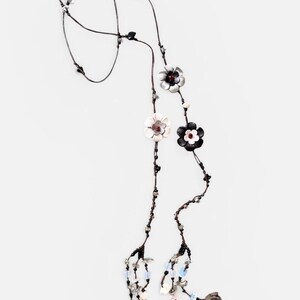May include: A long necklace featuring a thin dark cord adorned with multiple flower-shaped charms in black, white, and silver tones. The design includes small beads in clear, blue, and black colors, with dangling floral elements at the ends.