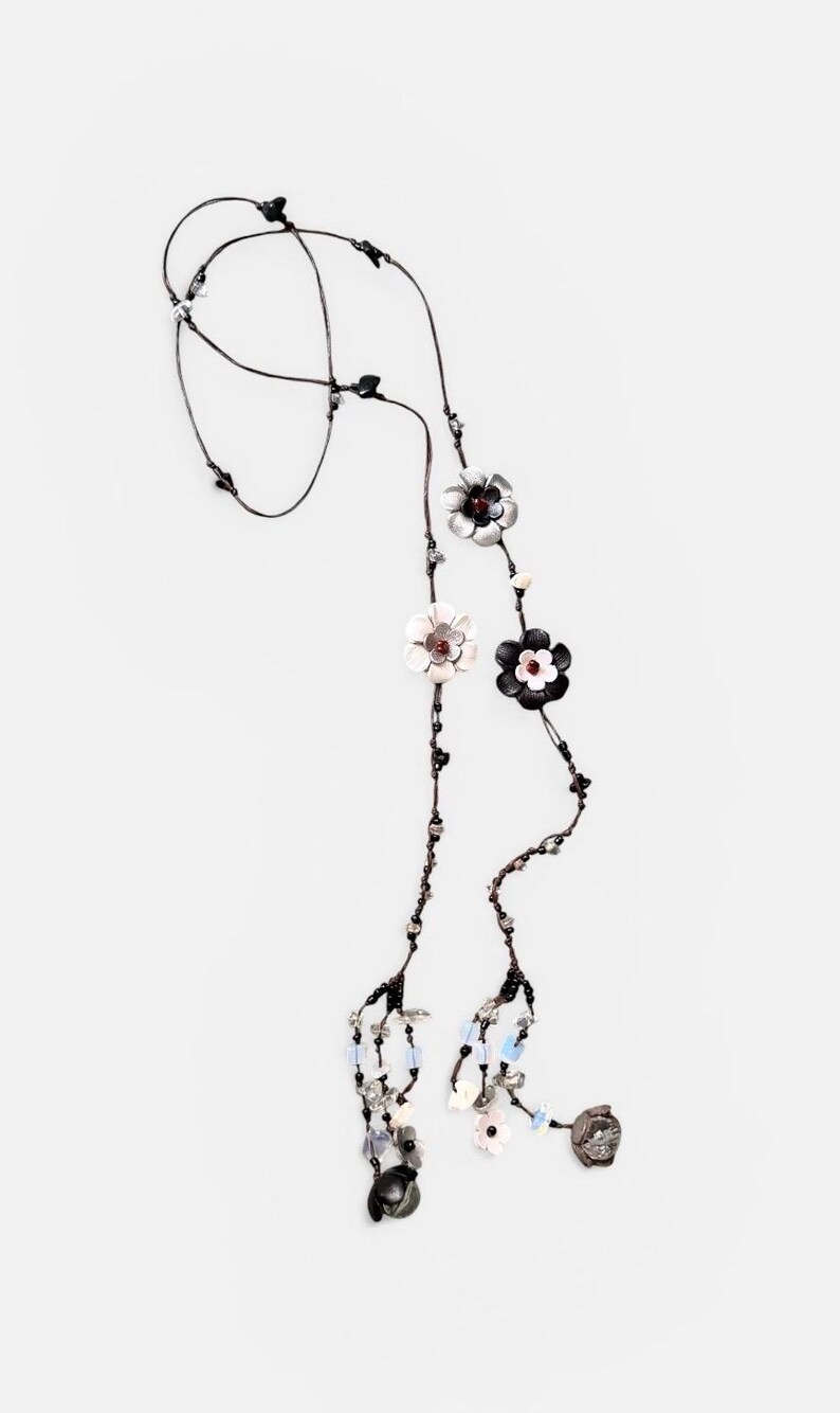 May include: A long necklace featuring a thin dark cord adorned with multiple flower-shaped charms in black, white, and silver tones. The design includes small beads in clear, blue, and black colors, with dangling floral elements at the ends.