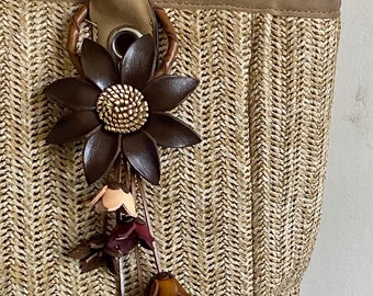 Könnte beinhalten: Eine gewebte Stroh-Tragetasche mit einem braunen Lederblumen-Anhänger. Der Anhänger hat eine große, dunkelbraune Blume mit goldenem Zentrum und kleinere, baumelnde Blumen in Braun- und Beigetönen. Die Tasche hat einen beigen Griff.