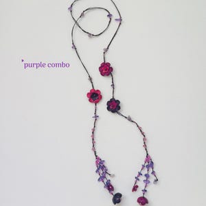 May include: A long necklace featuring a thin dark cord adorned with small purple and pink beads, several flower-shaped charms in shades of magenta and dark purple, and dangling beaded tassels at the ends. The text purple combo is visible.