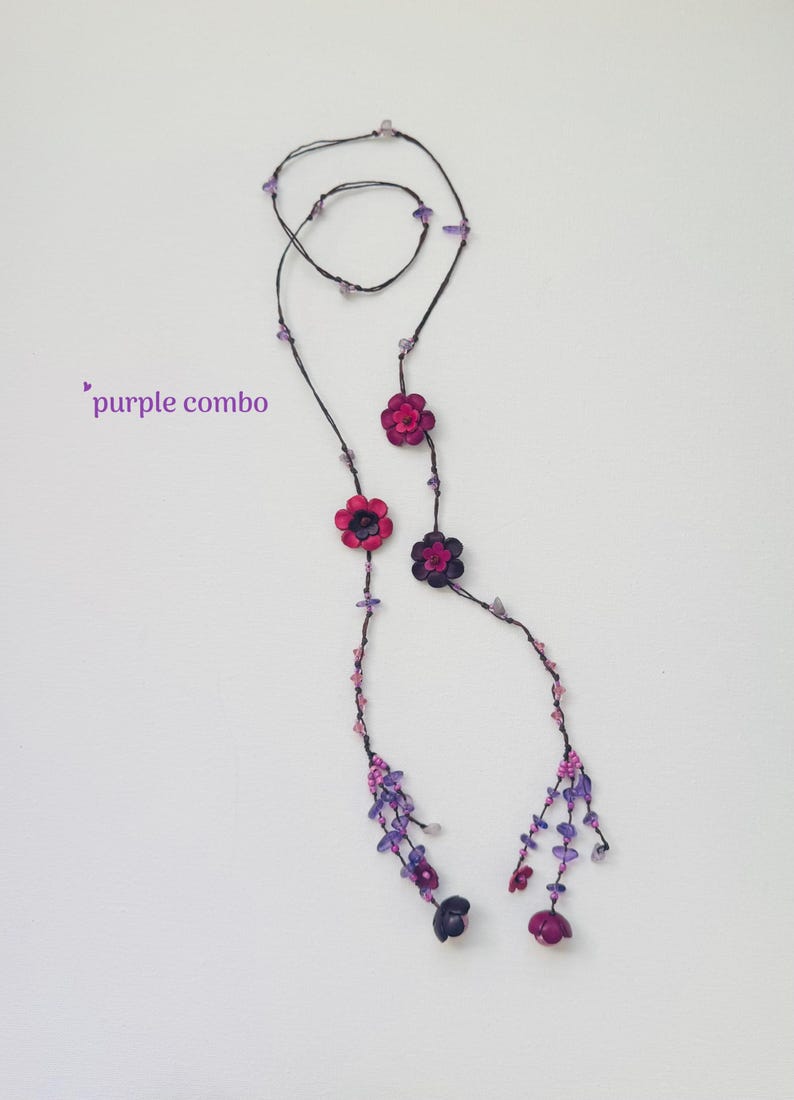 May include: A long necklace featuring a thin dark cord adorned with small purple and pink beads, several flower-shaped charms in shades of magenta and dark purple, and dangling beaded tassels at the ends. The text purple combo is visible.