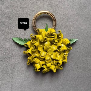 Op de afbeelding: Een gele leren bloem sleutelhanger met een goudkleurige ring. De bloem heeft gedetailleerde, gekrulde bloemblaadjes en groene bladeren. Het woord "GREEN" staat in een zwarte tekstballon.