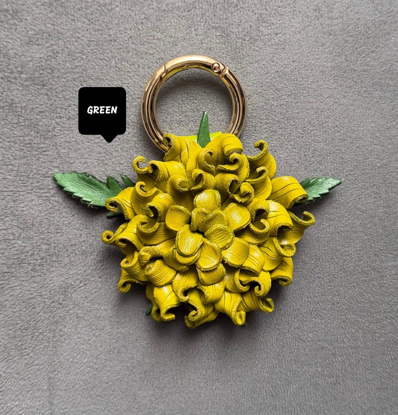 Op de afbeelding: Een gele leren bloem sleutelhanger met een goudkleurige ring. De bloem heeft gedetailleerde, gekrulde bloemblaadjes en groene bladeren. Het woord "GREEN" staat in een zwarte tekstballon.